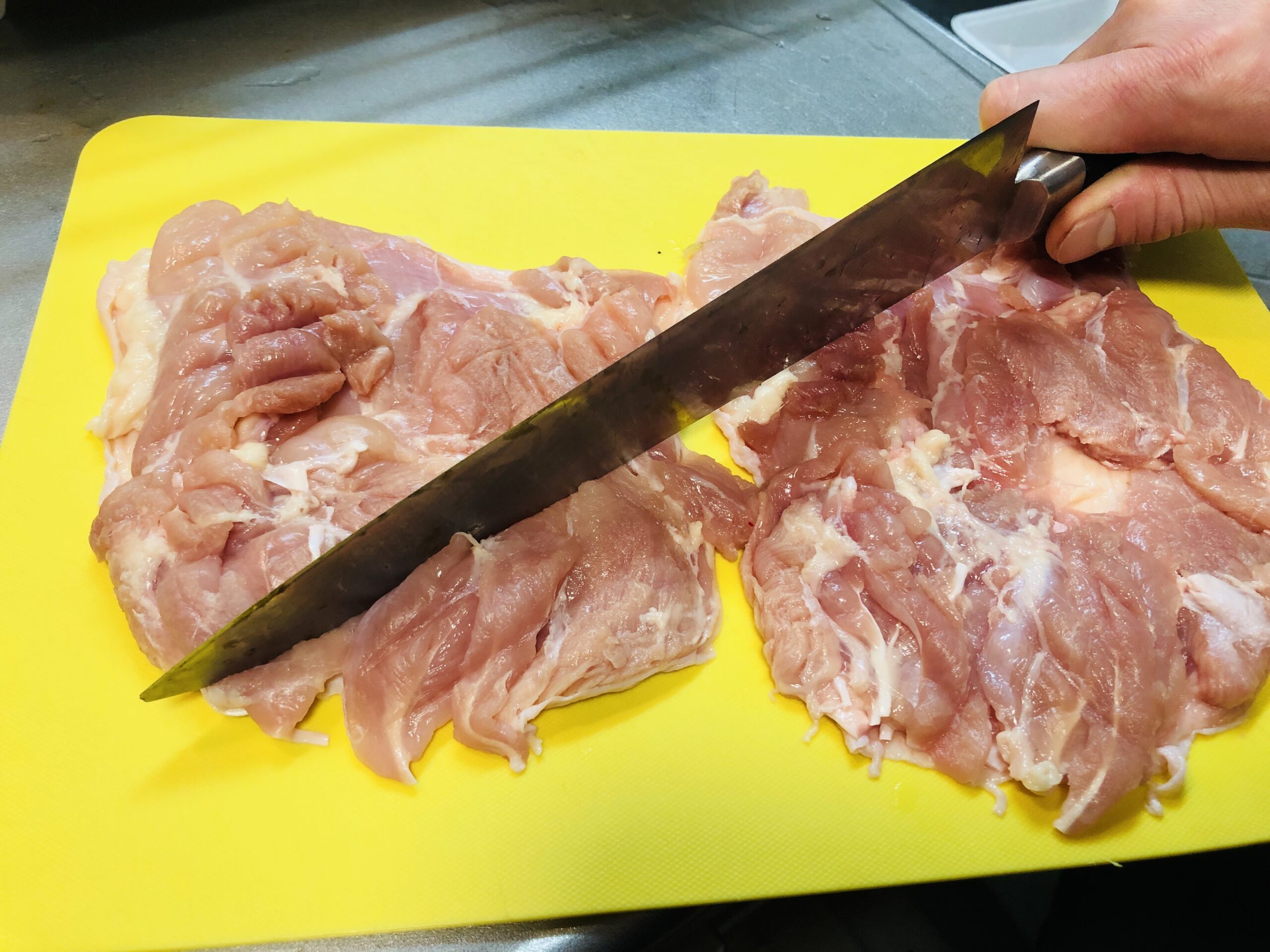 まな板の上で鶏もも肉を皮を下にしておいて、包丁の背で肉の厚い部分を叩いて均している様子の画像です